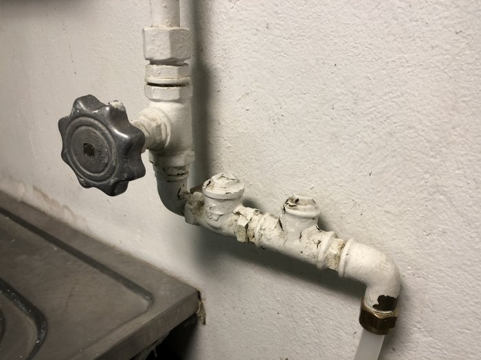 En bild på ett vitt vattenrör med en röd ventilknopp på vägg, med tecken på äldre lagningar och en öppen ände.
