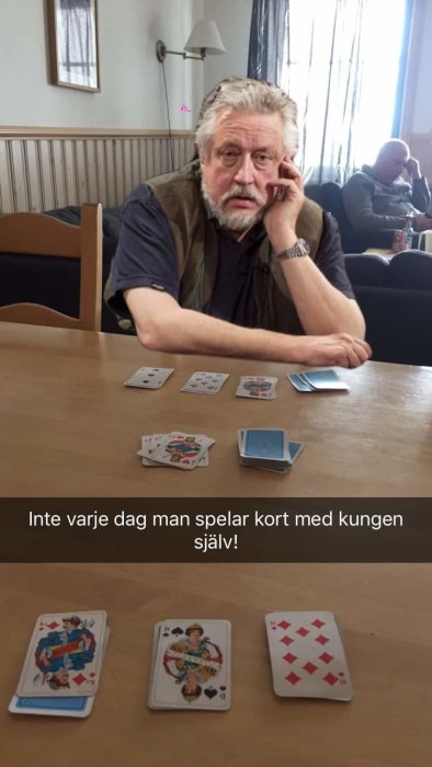 Man sitter vid bord spelar kort, uppvisade spelkort med text "Inte varje dag man spelar kort med kungen själv!