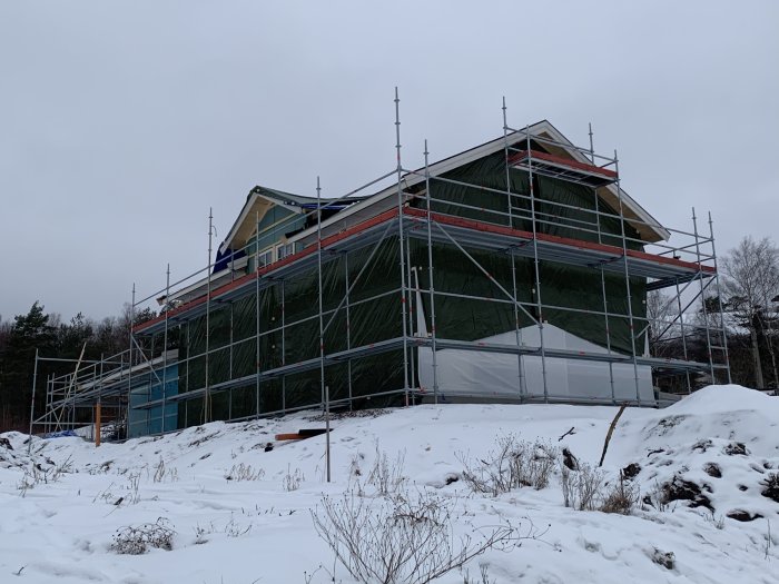 Hus under konstruktion täckt med presenning och byggnadsställningar på en snöig tomt.