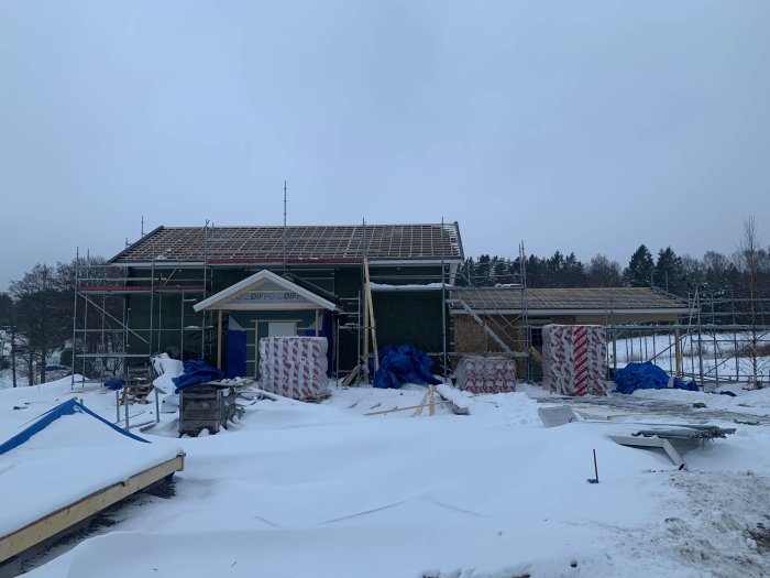 Hus under konstruktion med OSB på vägg mot garaget, täckt med presenning, i snöigt landskap.