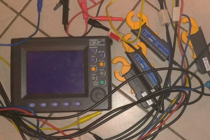 En digital mätutrustning blandat med flera trassliga kablar och klämmor på ett golv.