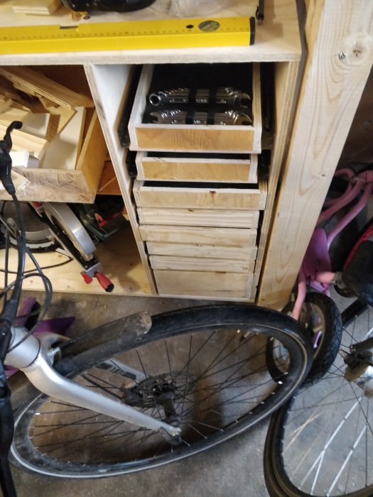 Mätning av träbitar för att skapa lådfronter i ett rörigt garage med synliga verktyg och cykeldelar.