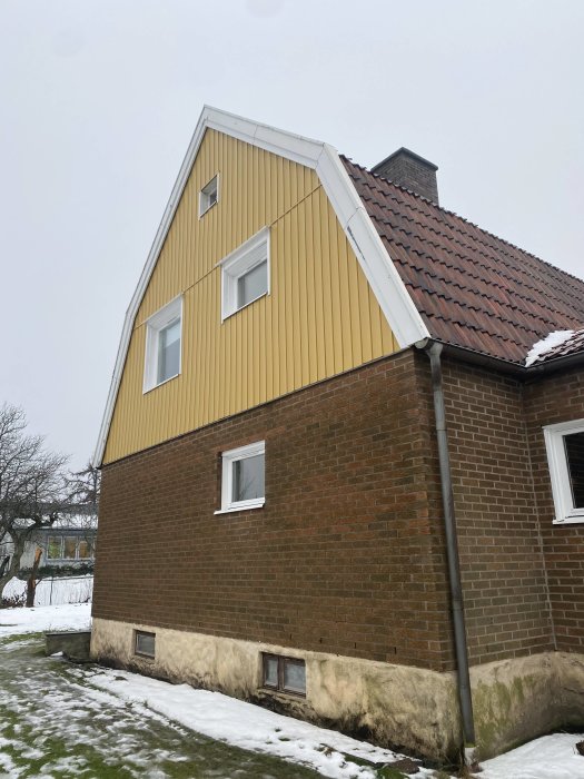 Husfasad med tegel på nedervåningen och gul träpanel på övervåningen, fönstren sitter djupt i väggen.