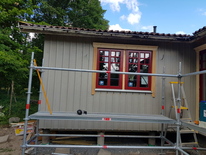 Del av husfasad under renovering med röda fönsterfoder och ställning framför.