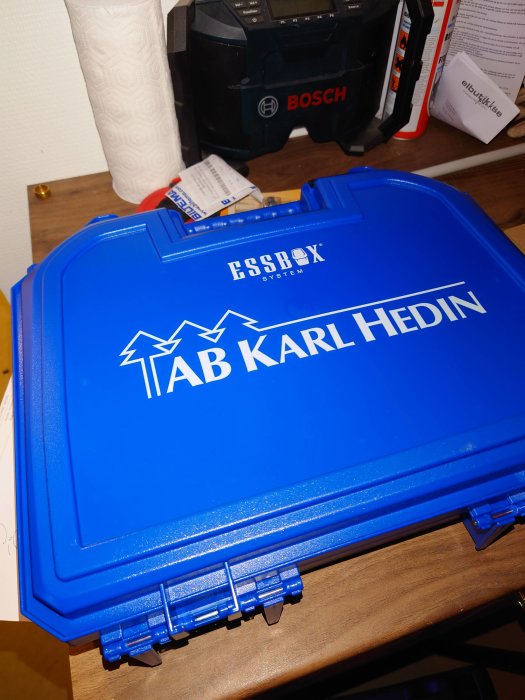 Blå Essbox skruvsorteringslåda med texten 'TAB KARL HEDIN' på en arbetsbänk, bredvid verktyg och tillbehör.