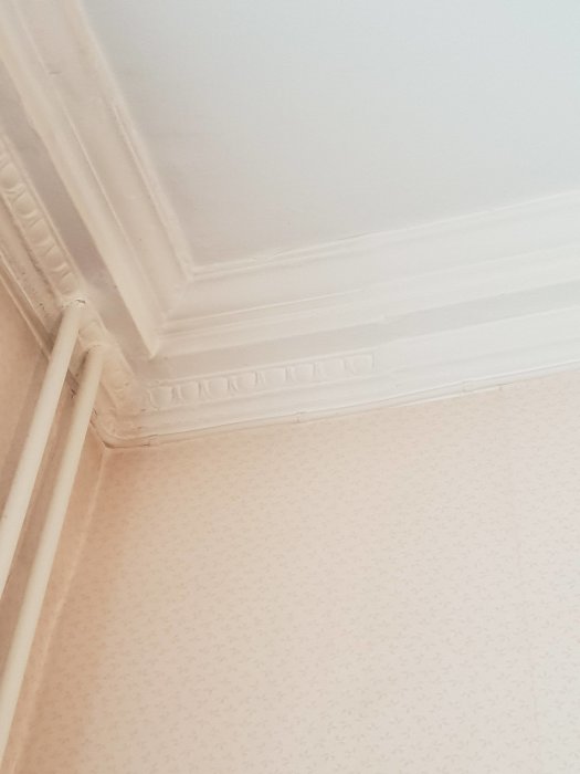 Hörnan av ett rum med tapeter, där ursprunglig stuckatur är delvis dold under ett lager spackel eller puts.