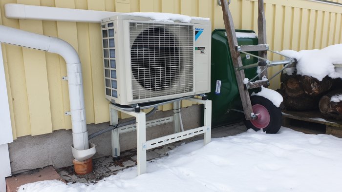 Värmepump monterad på ställning med snö, avloppsrör och dagvattenavledning vid husvägg.