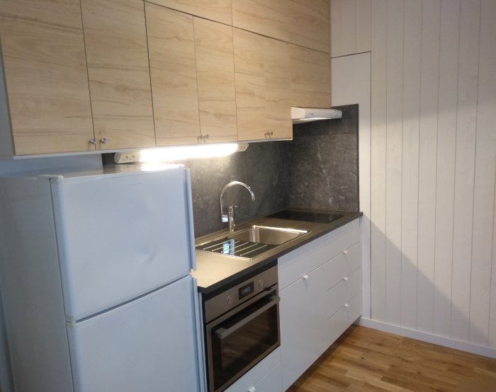 Nyligen renoverat kök med vita skåp och trädetaljer, höga väggskåp och en buckla på kyl/frysens dörr.