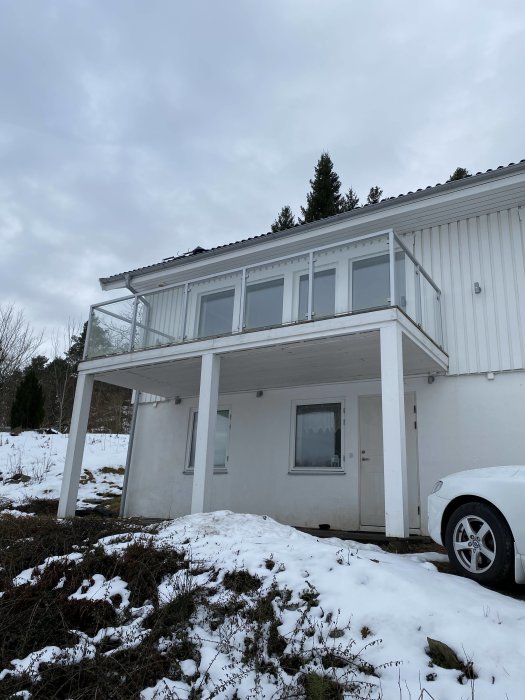 Vit tvåvåningshusfasad med balkong och snö på marken, delvis molnhimmel och en bil till höger.