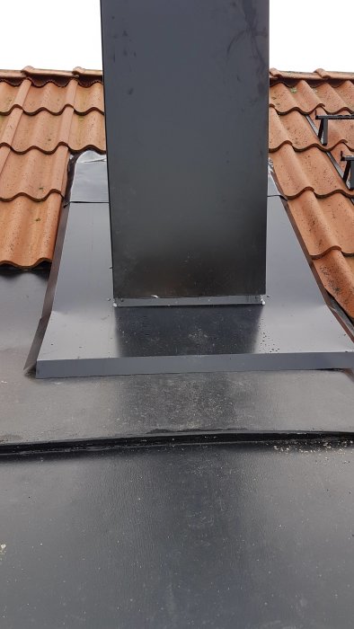 Takdetalj där en svart skorsten möter platt takbeklädnad med tegelpannor i bakgrunden.