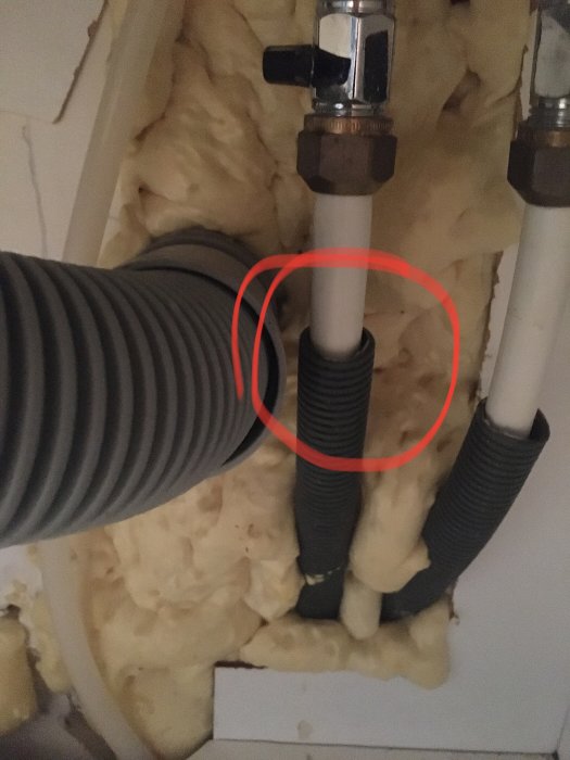Mörkt plaströr markerat med röd cirkel som läcker vatten, omgivet av isolering och andra rör.