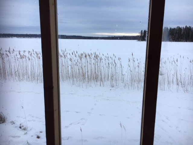 Vy genom fönster av snöklädd sjö med vass och fotspår i förgrunden och molnigt himlavalv ovan.