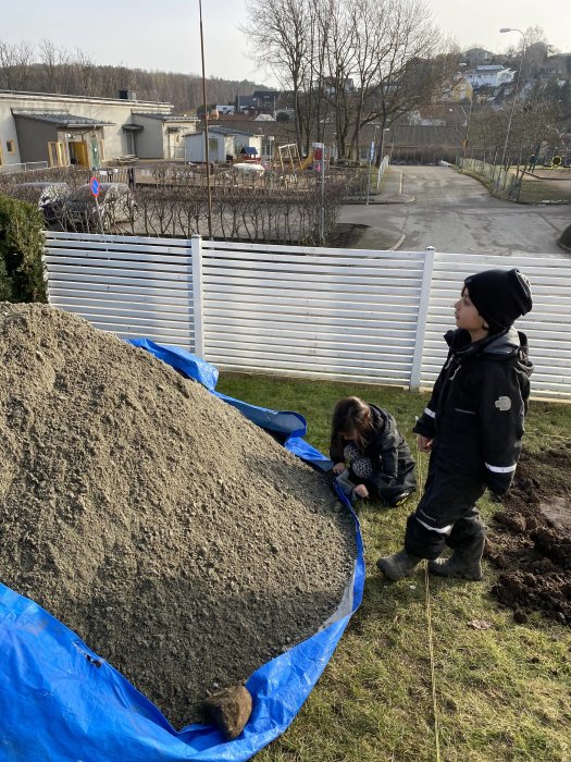 Två barn vid en stor hög grus på en blå presenning i en trädgård, med lekplats och bostadshus i bakgrunden.