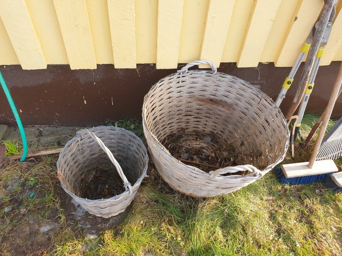 Två tomma flätade trädgårdskorgar, beskärningssax och sopkvast vid en gul husvägg.