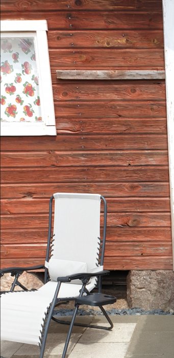 Rödmålad trävägg på uthus med falu rödfärg, intill en vit dyna på en svart solstol, över grus och stenplattor.