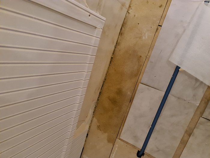 Golvrivning med synlig fuktfläck på spånskivebit nära en vit vägg och kakel, delvis täckt med ett nytt golv.