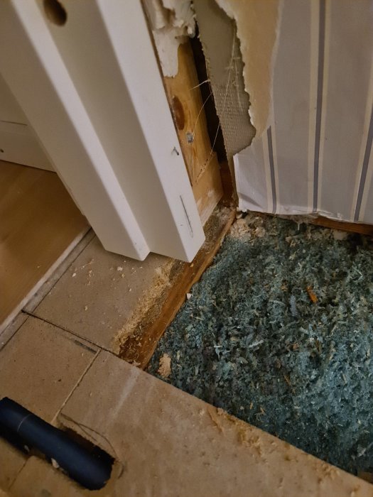 Borttagen del av golvspån vid dörrkarm, exponering av underliggande regel och isolering, redo för reparation.