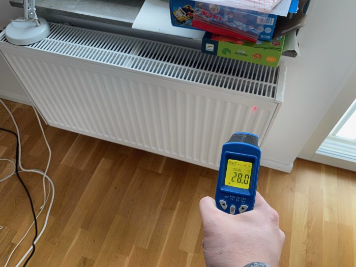 Hand håller en värmekamera som visar 20.8°C framför ett radiator element i ett rum.
