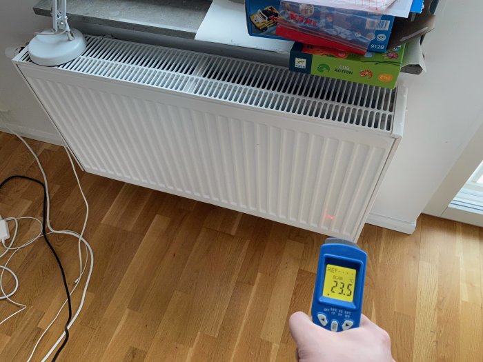 Hand håller en infraröd termometer riktad mot ett element, visar 23.5°C, elementet ser kallt ut.