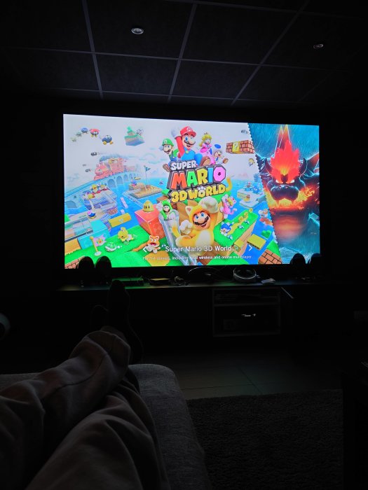 Person spelar Super Mario 3D World på TV i mörkt rum med benen upp på en ottoman.