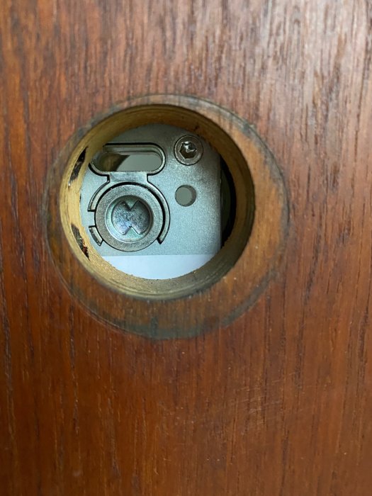 Närbild av ett låshus inuti en dörr, synligt genom rund urfräsning i brunt trä.
