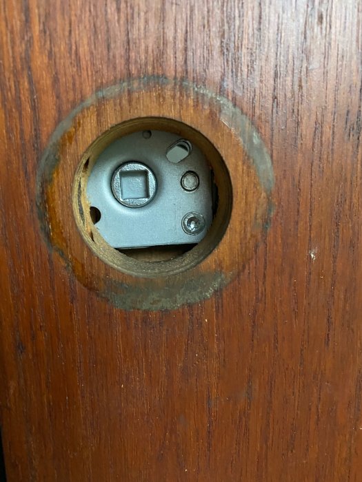 Utskuret hål i en brun dörr för ett låshus, med synliga spår av bearbetning och gammal inramning.