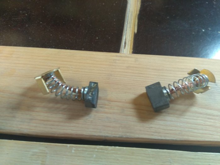 Två utslitna kolfiberborstar med fjädrar och kontaktklämmor på ett träbord.