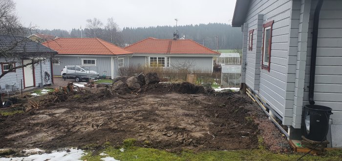 En grävd och ojämn trädgård med stora stenar vid hus med röda tak, planerad för att bygga stödmur.