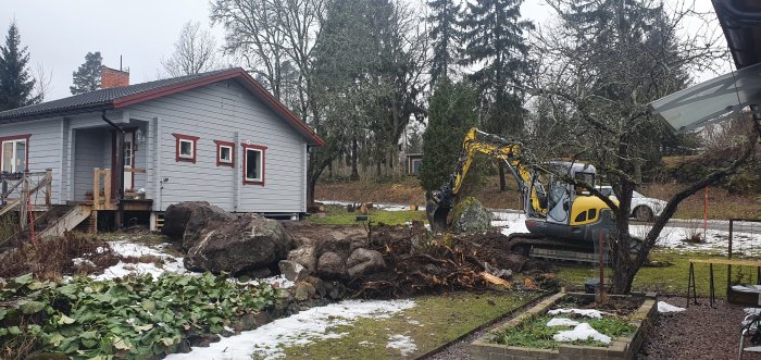 Bild på grävmaskin och stora stenblock vid sluttande trädgård nära ett hus.