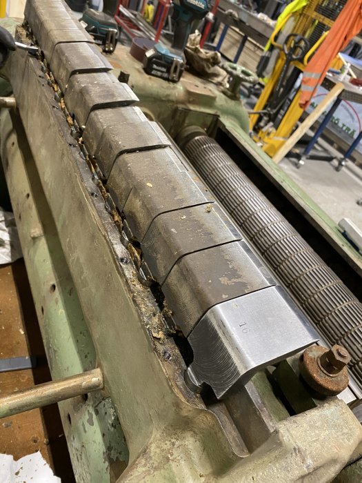 Metalldel med fjädrande "knivar" som tros vara ett bakslagskydd, placerade på en maskindel i en verkstad.