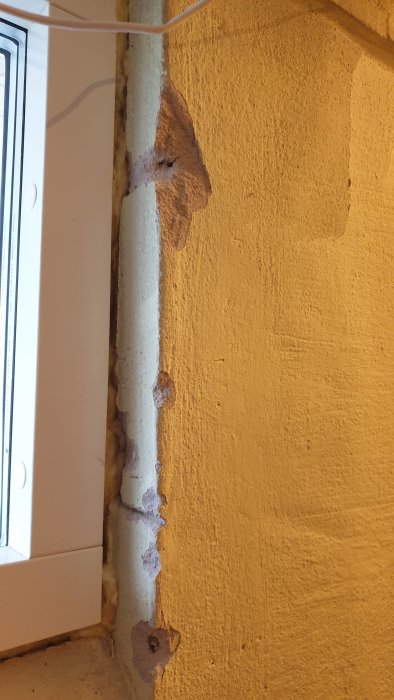 Skadad gul källarvägg med lossad puts runt ett fönster och exponerad underliggande struktur.