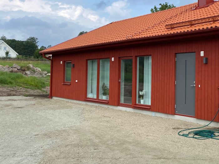 Röd trävilla med nytt grusunderlag förberett för trädäck, grov bergkross nära huset och slang på marken.