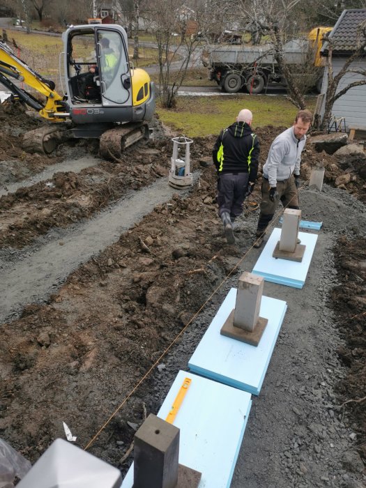 Två personer som arbetar med utplacerade plintar på en byggarbetsplats med en grävare i bakgrunden.