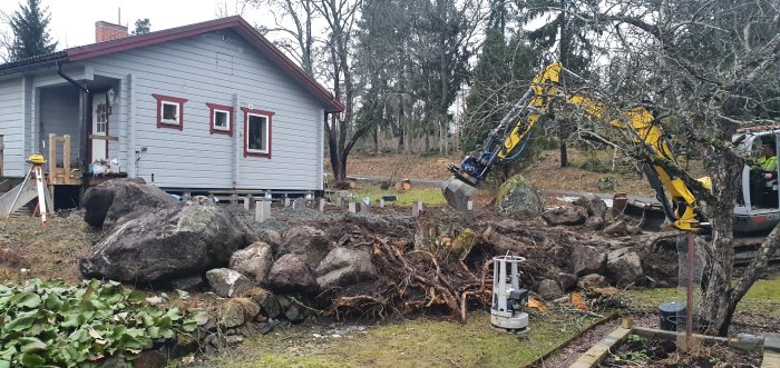 Grävmaskin i arbete nära hus med stora stenar och nyligen placerade betongplintar i en trädgård.