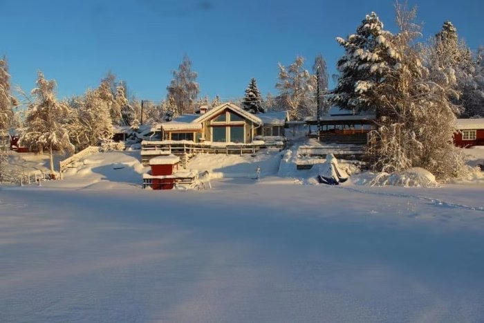 Vinterbild av en sommarstuga från 50-talet, påbyggd, med snötäckt sadeltak och omgivning.