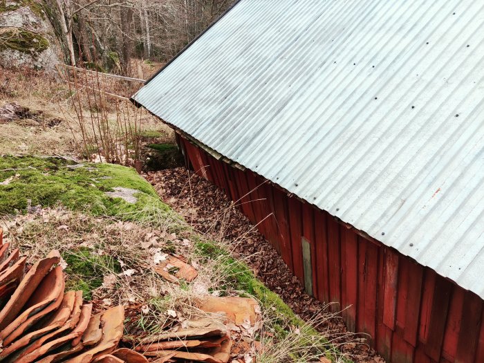 Röd vedbod med skadad hörnstolpe vid sluttning och berg, vattenskador synliga.