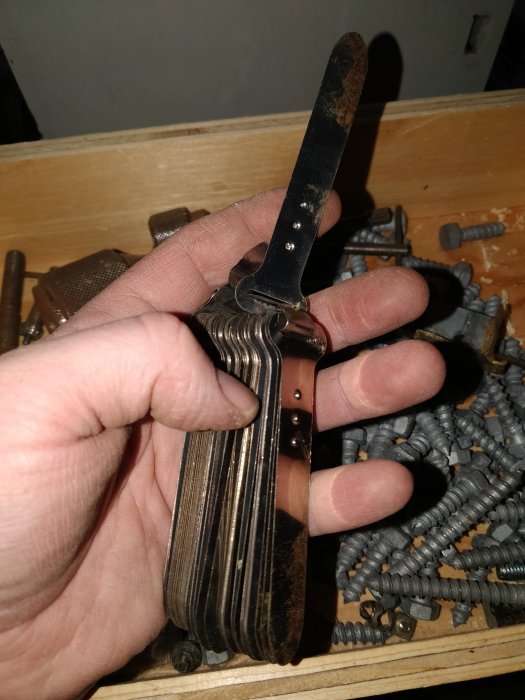 Hand håller ett gammalt multiverktyg med utfällda blad över en låda med skruvar.