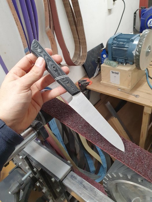 Hand håller kniv med slipslipad yta och trähandtag i verkstad med slipband i bakgrunden.