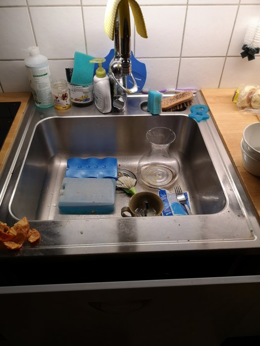 Köksdiskbänk med disk, tvåldispenser, svampar, och borste nära en öppen vattenkran.