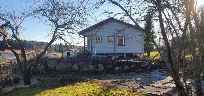 Nygrävd mark och stödmur av stora stenar framför ett vitt hus med veranda och röda fönsterkarmar.