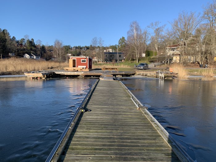 Soligt landskap med halvfrusen sjö, brygga och röd stuga, indikerar en start på vår med bastu- och vakbadstraditioner.