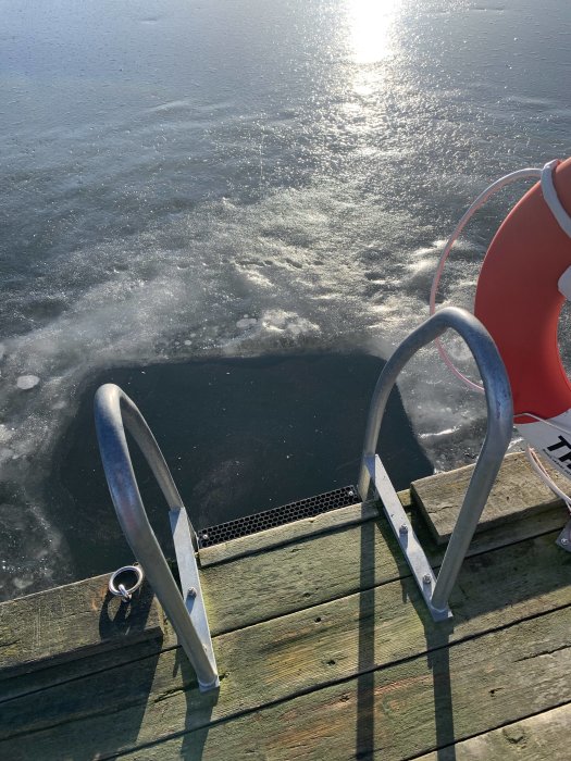 Solreflektioner på fruset vatten sedd från en träbrygga med stege och livboj i solljus.