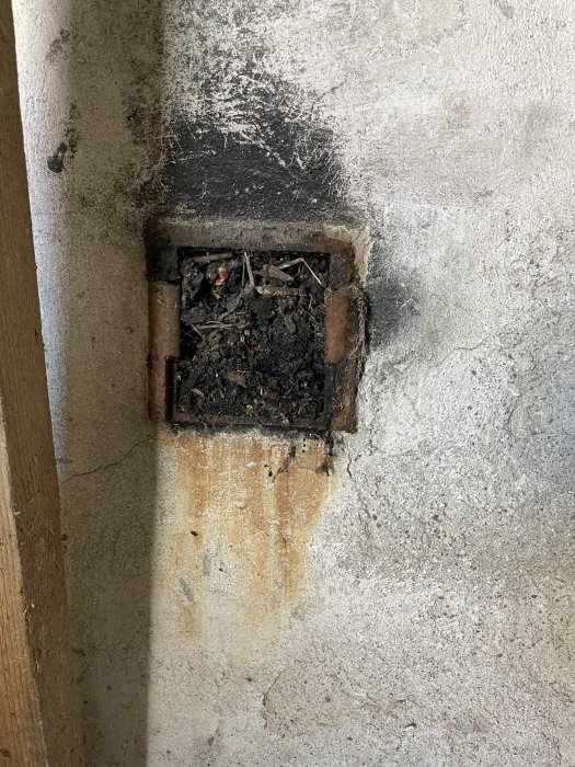 En gammal bakugns lucka med sot och aska i en sliten betongvägg, tecken på historisk användning.