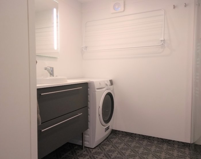 Renoverat badrum med grå kommod, tvättmaskin, ljus inredning, och väggmonterad torkställning.