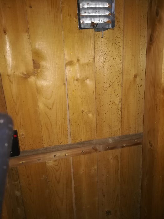 Trävägg i vind med svarta prickar runt en ventilationslucka, indikerar möjlig fukt- eller mögelskada.