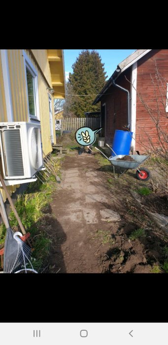 Kortsida av hus med gul färg, sorglig stenrad och jord, ogräs, skottkärra och luftvärmepump syns.