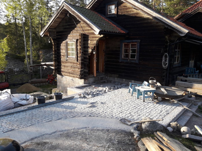 Anlagd kullerstensuppfart framför en timmerstuga under renovering, med byggmaterial utspritt.