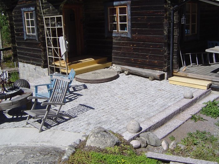 Traditionellt mörkt timmerhus med kullerstensbelagd gård, trädgårdsmöbler och nybyggd trätrappa.