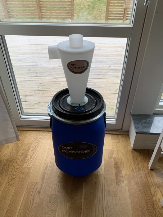 En Dust Commander P130 dammavskiljare i vit och blå färg står på ett trägolv vid ett fönster.