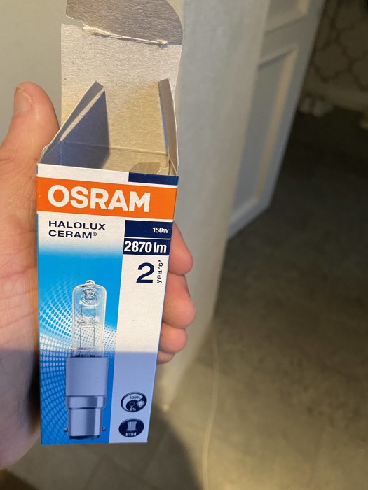 Hand håller en Osram Halolux Ceram 150W halogenlampa i förpackning.